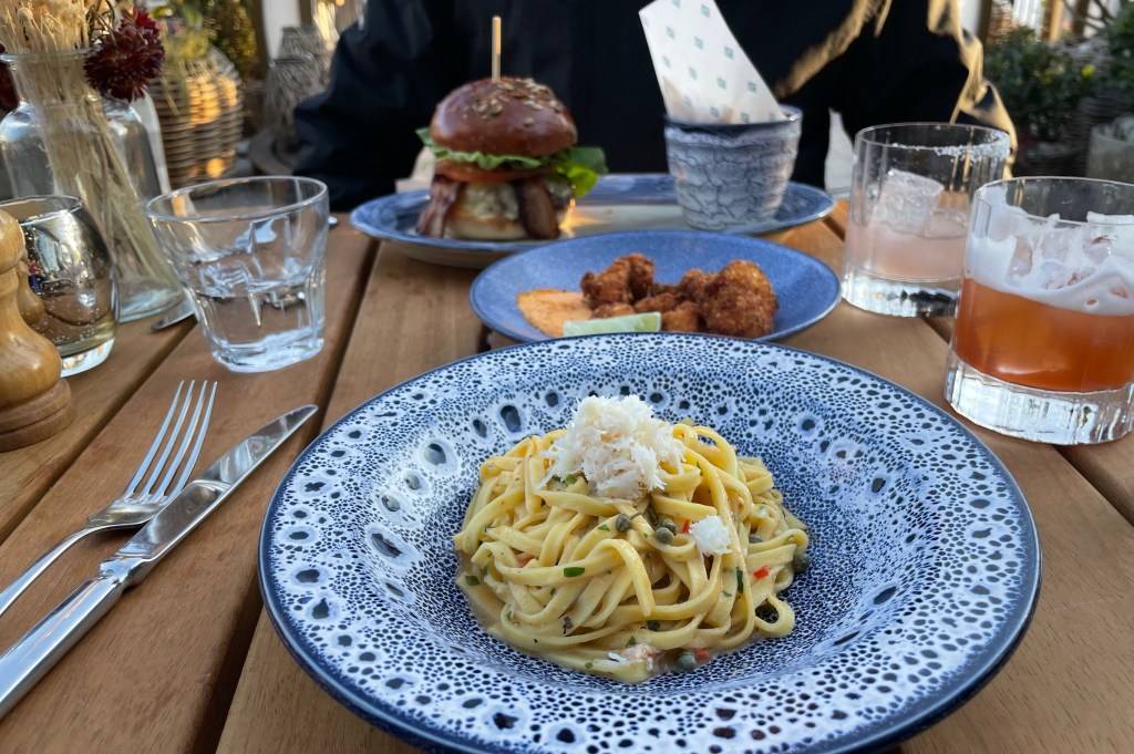 A plate of crab linguine in the foreground next to a glass of Coppa Sour, with a plate of fried chicken and dipping sauce,  a glass of Hibiscus Martini and a plate with a Coppa burger and skinny fries in the background.