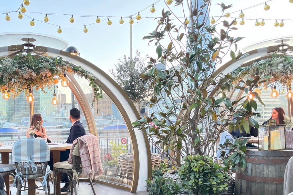 A couple sits in a glass igloo with their heads turned facing toward the river behind them.  There is a light fixture decorated with a plant Garland hanging from the top of the igloo,  and one of two empty chairs opposite the couple is draped in a blanket. Next to the igloo is a plant in a pot, and another occupied igloo. Above it all is a network of hanging lights.