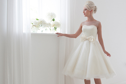 A model is wearing the Daisy dress by House of Mooshki. It is white, knee-length, and covered in polkadot lace. It has a sash with a bow on the front.